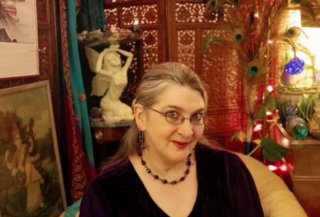 A gently smiling woman wearing dark purple sits before a carved teak screen from India and a statue of a kneeling fairy. A picture of the Hindu goddess Sarasvati appears next to her, on the viewer's left. Pink lights twinkle next to peacock feathers on the viewer's right.